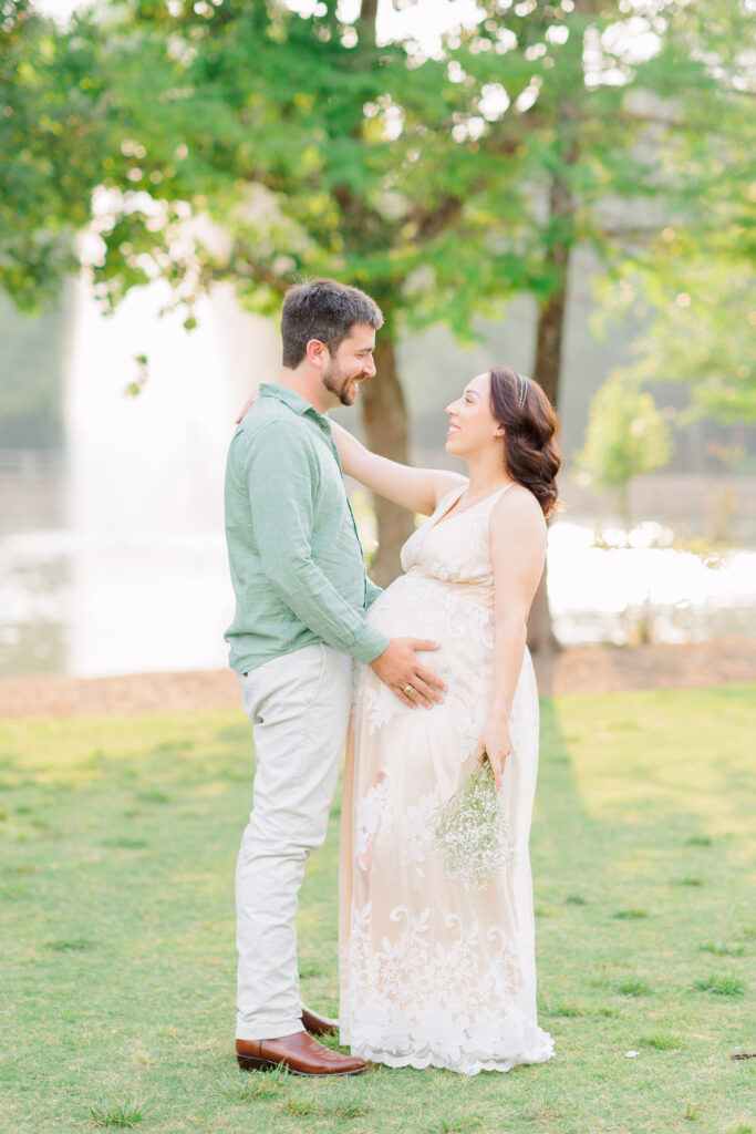 couple poses together with baby bump in front of a water fountain at rob fleming park for Cypress Maternity Photographer allie nichols photography