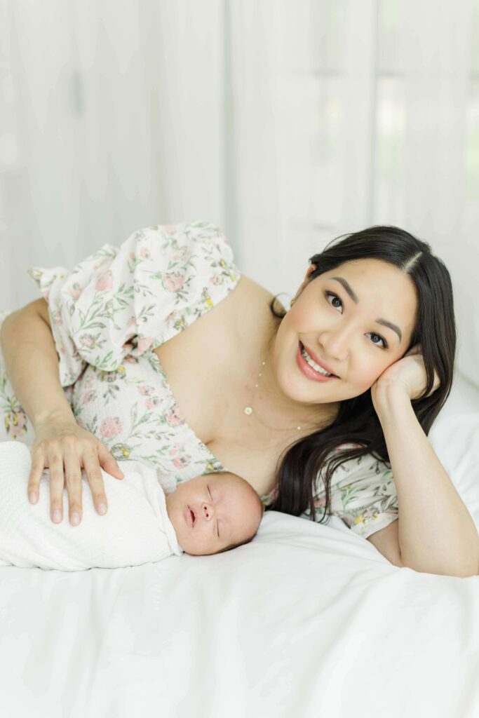 Mom smiles at the camera side lying with baby on a white bed in a houston studio