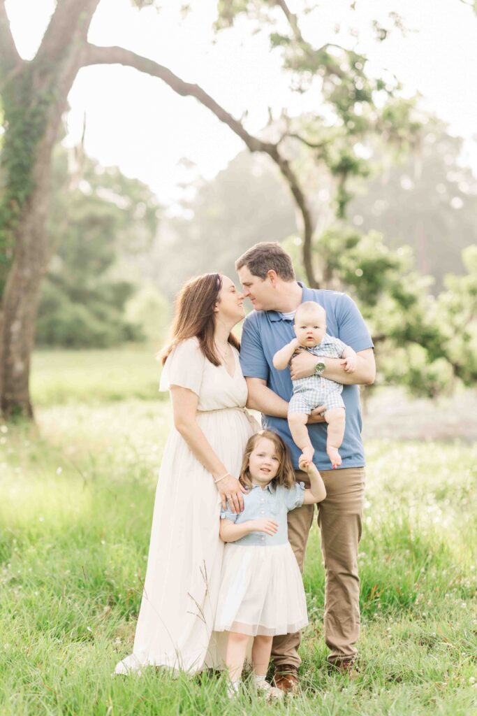 mom and dad nuzzle noses together holding baby boy and big sister in front of a mossy tree