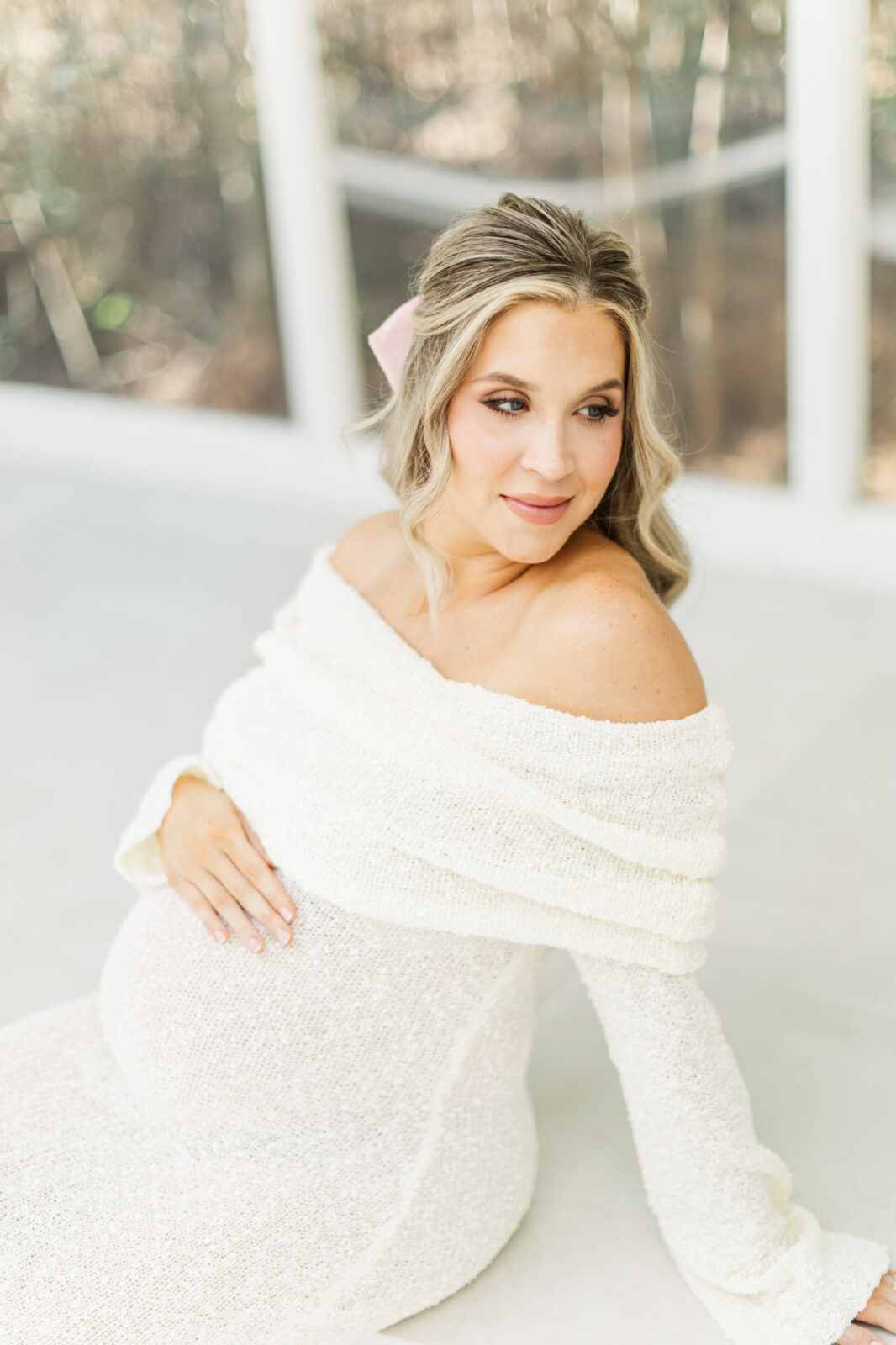 Dreamy indoor maternity photo featuring soft white tones and a mom embracing her baby bump near a window.