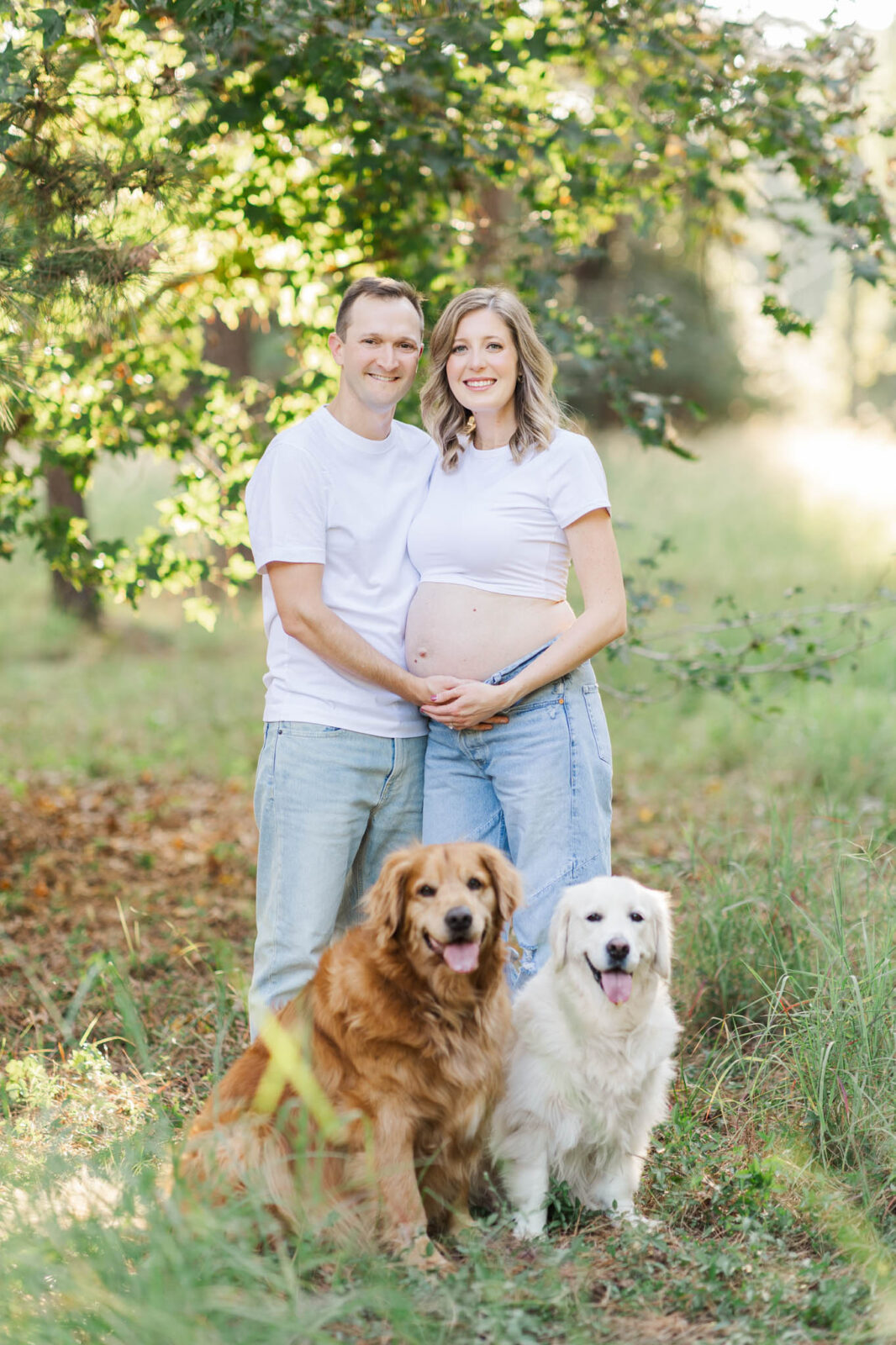 Husband embracing his pregnant wife from behind as she holds her baby bump in front of their dogs at East End Park In Kingwood