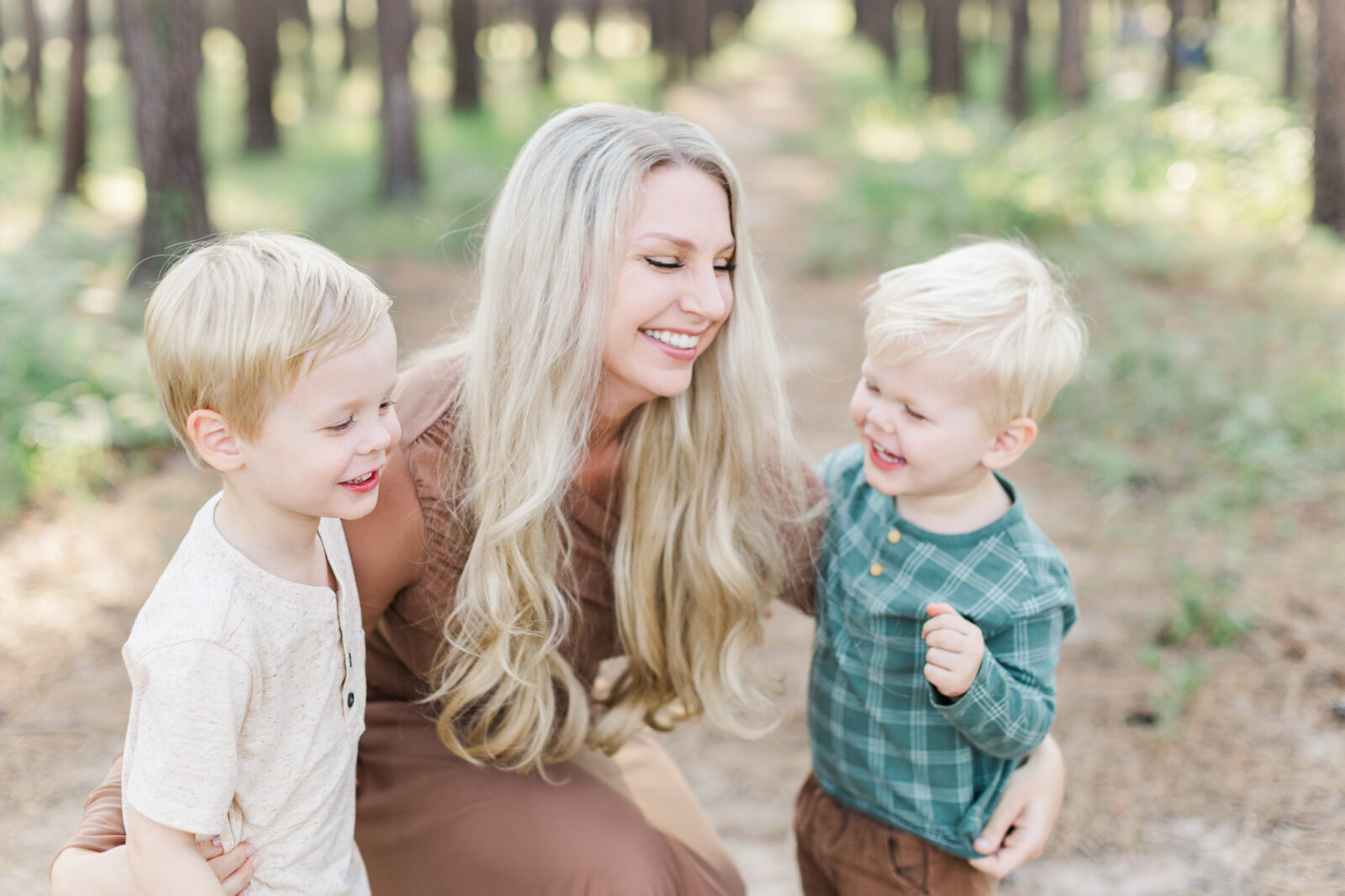 Expecting mother smiling while her sons stand beside her under tall pine trees during a Houston maternity photoshoot.
