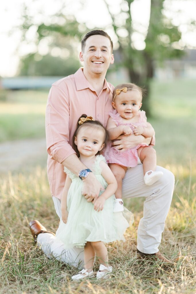 dad kneels to hold two daughters under softly lit tree wearing boutique outfits